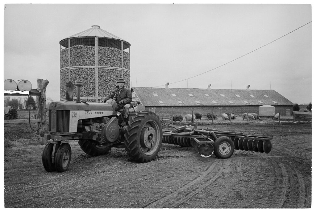 Man driving a John Deere tractor, pigs and corn crib in the background