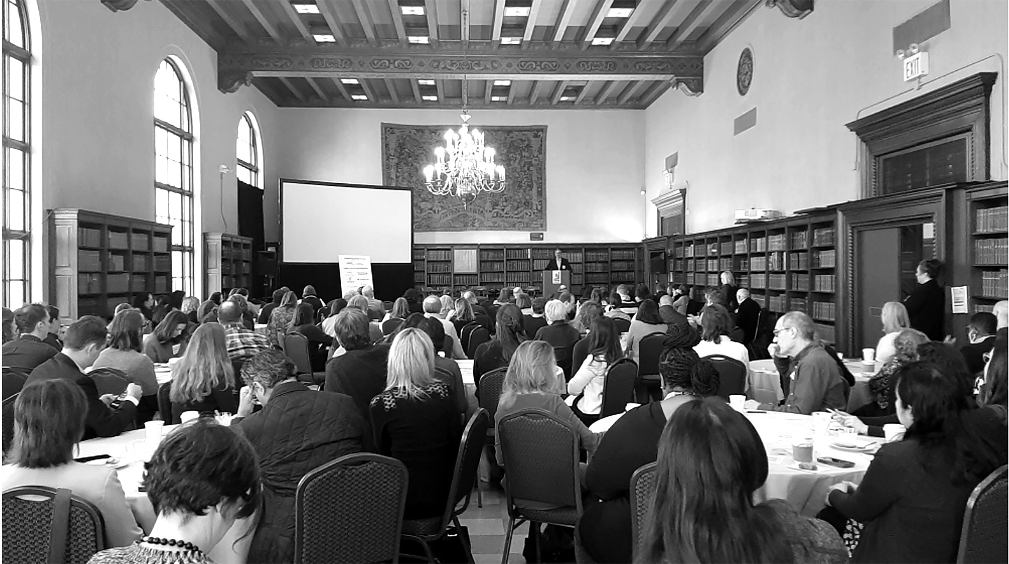 Black and white image of people gathered in a library room, facing speaker at podium