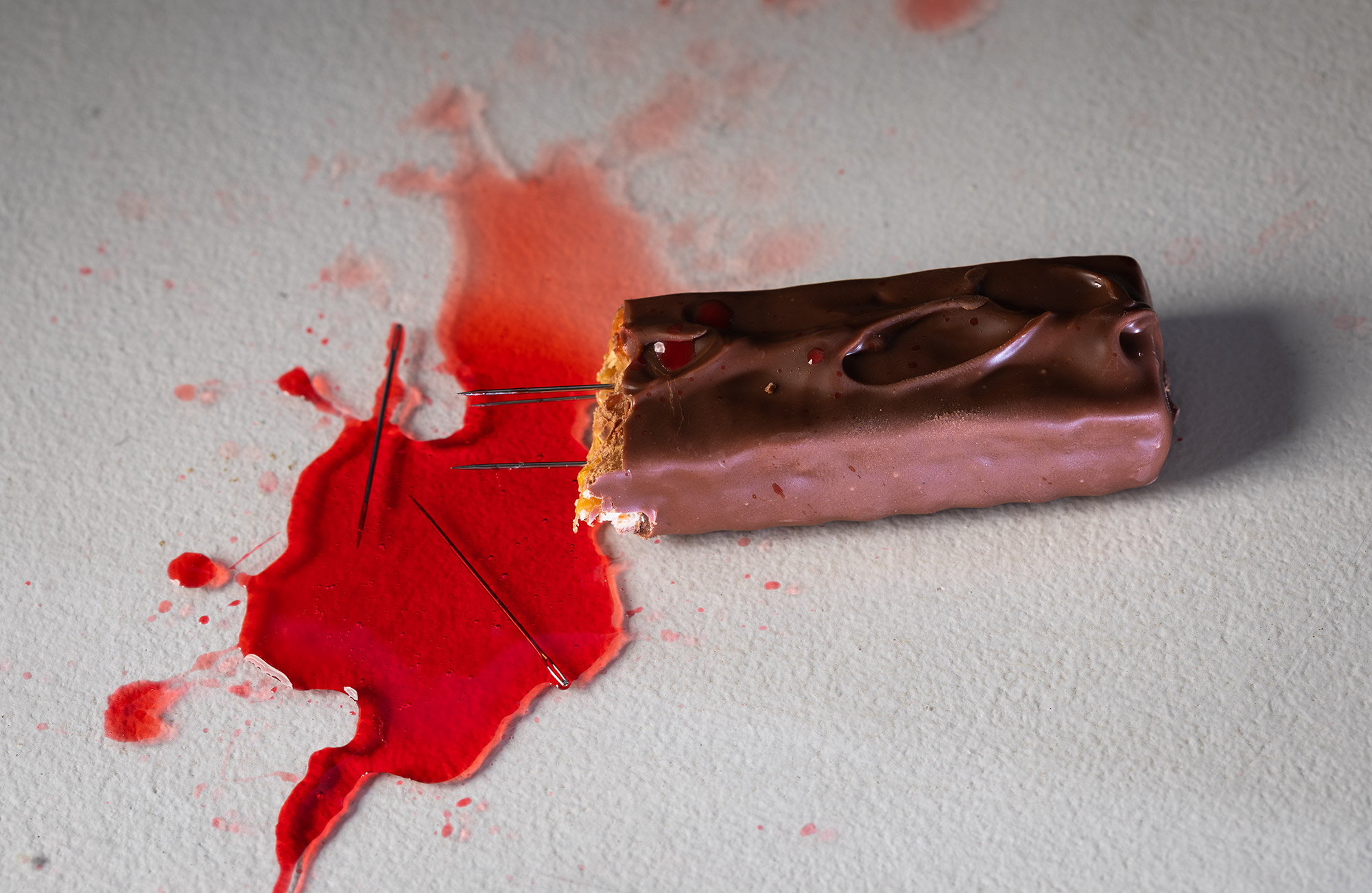 Chocolate bar with needles sticking out, in a pool of red liquid (blood)