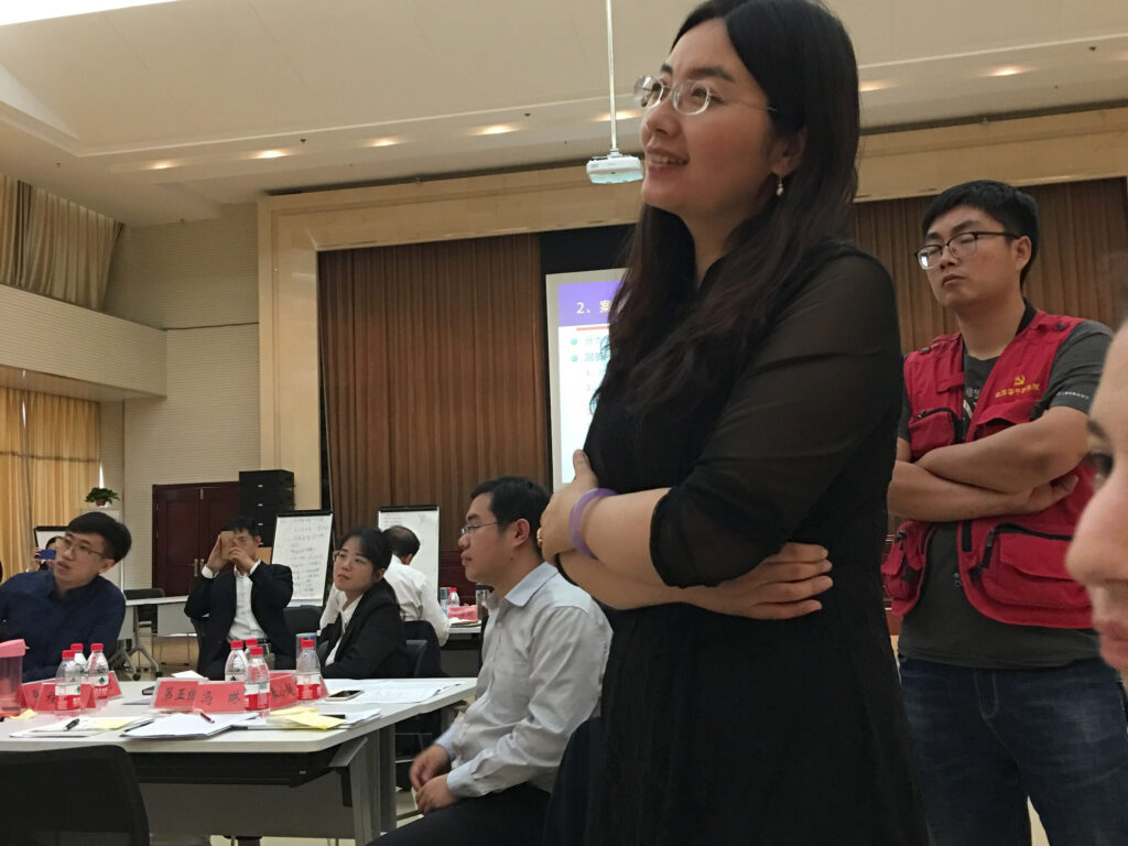 Photo of woman with glasses wearing a black dress listening to academic presentation, with other seated colleagues at tables in background