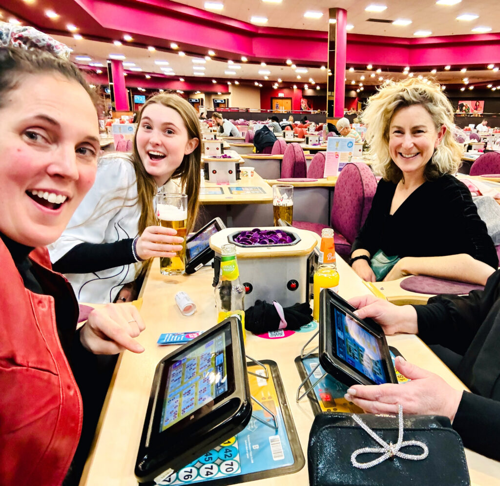 A selfie-style photograph of three women smiling and laughing at a table in a large, brightly lit bingo hall. A fourth woman's hands are visible. They have drinks, paper bingo cards, and electronic bingo tablets set up in front of them. The woman on the left is pointing excitedly at her screen. The background shows rows of tables with purple chairs and other patrons playing under a distinctive pink ceiling.
