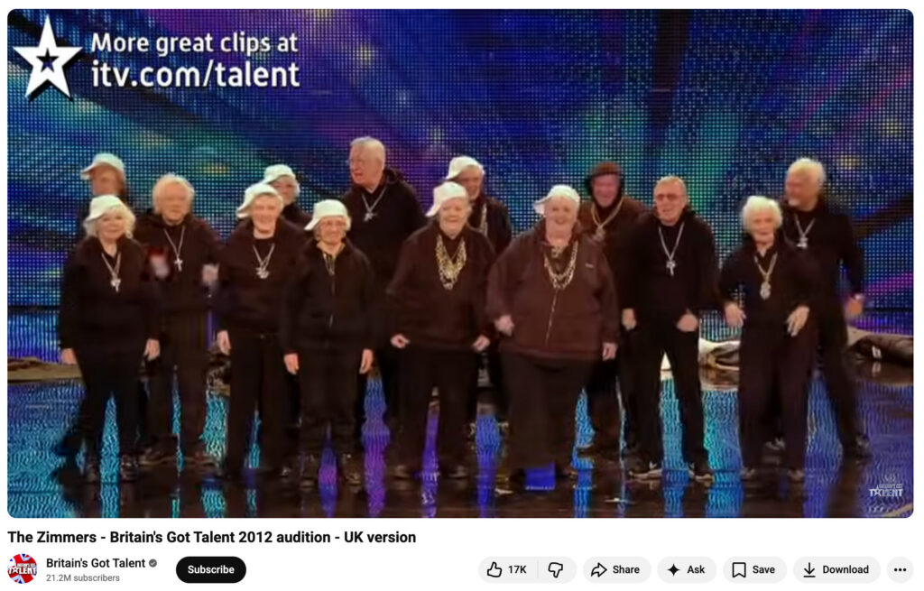There are 14 older men and women in the image, all standing on a stage, mostly wearing black tops and pants, and fashioning large, noticeable necklaces. Many of the women are also wearing white baseball caps. The screenshot is from a YouTube video of the 2012 Britain's Got Talent audition. 