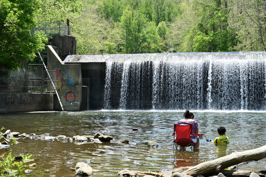 woman and child sitting in river, looking up at spill-over dam, with additional infrastructure on left