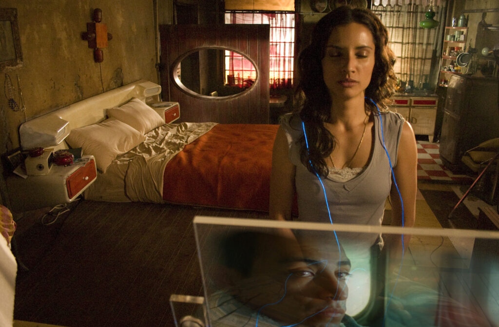 Film still from Sleep Dealer showing dark haired woman named Luz sitting at a laptop. The face of Memo is reflected in the back of the laptop.