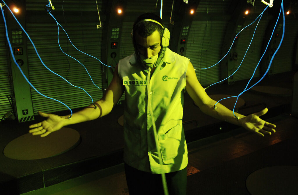 Film still from Sleep Dealer showing Memo with arms outspread and blue cables coming out of his arms. He is lit in a green light.